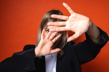 Beyaz tişörtlü genç bir kadın parlak turuncu bir arka plan karşısında elleriyle yüzünü kapatıyor. Korku, mahremiyet, reddedilme ya da sosyal endişe kavramı