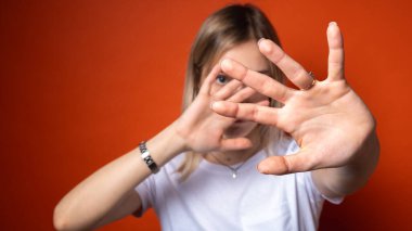 Beyaz tişörtlü genç bir kadın parlak turuncu bir arka plan karşısında elleriyle yüzünü kapatıyor. Korku, mahremiyet, reddedilme ya da sosyal endişe kavramı