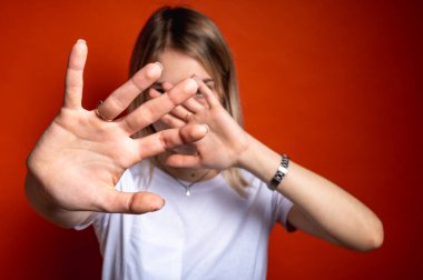 Beyaz tişörtlü genç bir kadın parlak turuncu bir arka plan karşısında elleriyle yüzünü kapatıyor. Korku, mahremiyet, reddedilme ya da sosyal endişe kavramı