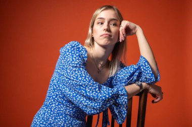 Kendine güvenen, mavi çiçekli elbiseli genç kız turuncu arka planda sandalyeye yaslanıyor. Havalı, anlamlı ve modern moda portresi ruh hali, duygular, stres, yansımalar