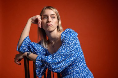 Kendine güvenen, mavi çiçekli elbiseli genç kız turuncu arka planda sandalyeye yaslanıyor. Havalı, anlamlı ve modern moda portresi ruh hali, duygular, stres, yansımalar