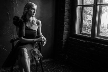 Siyah beyaz fotoğraf, siyah elbiseli zarif genç kız klasik deri koltukta oturuyor, kendine güven ve zarafet saçıyor. Karanlık bir geçmişi olan şık bir portre, seçici bir odak noktası.