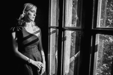 Siyah beyaz fotoğraf, siyah gece elbisesi içinde zarif bir genç kadın eski ahşap pencerenin yanında dikiliyor ve düşünceli bir şekilde dışarı bakıyor. Yumuşak güneş ışığı, klasik atmosfer, romantik ruh hali