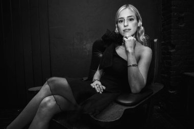 Siyah beyaz fotoğraf, siyah elbiseli zarif genç kız klasik deri koltukta oturuyor, kendine güven ve zarafet saçıyor. Karanlık bir geçmişi olan şık bir portre, seçici bir odak noktası.