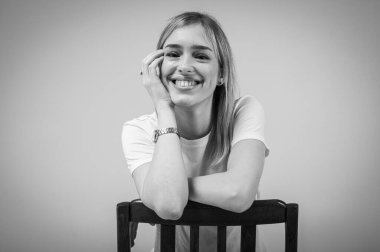 Siyah beyaz portre, mutlu, duygusal genç kız sarı saçlı gülümseyen ve tarafsız bir arkaplanda tahta bir sandalyeye yaslanan. Neşeli kadın, güven, neşe, pozitiflik, yazıtların yeri