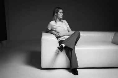 Beyaz deri bir kanepede oturan, beyaz t-shirt giyen, düşünen, kendinden emin genç bir kadının siyah beyaz fotoğrafı. Tarafsız bir geçmişi olan bir kadının modern minimalist portresi iş ve yaşam tarzı için mükemmel.