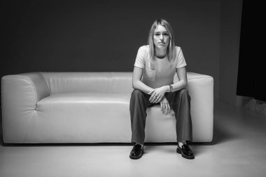 Beyaz deri bir kanepede oturan, beyaz t-shirt giyen, düşünen, kendinden emin genç bir kadının siyah beyaz fotoğrafı. Tarafsız bir geçmişi olan bir kadının modern minimalist portresi iş ve yaşam tarzı için mükemmel.