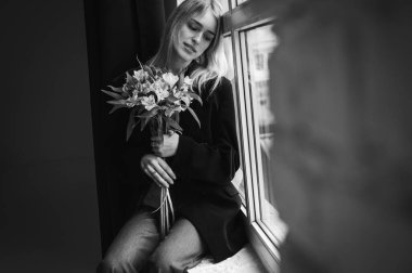 Siyah beyaz fotoğraf, siyah ceketli zarif, duygusal kız arka planda oturuyor ve odak noktasında güzel bahar çiçekleri var. Modern estetik ile stil, kompozisyon, seçici odaklanma