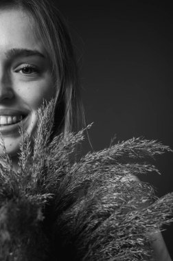 Siyah beyaz fotoğraf, beyaz tişörtlü genç bir kadın elinde bir buket kurutulmuş pampa otuyla turuncu arka planda sakin bir ifadeyle aşağıya bakıyor. Kırsal estetik ve doğal güzellik