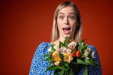 Mavi çiçekli elbisesiyle gülümseyen, güzel bir genç kız elinde parlak turuncu bir arka planda poz veren bir buket çiçek tutuyor. Bahar havası, doğal güzellik, neşeli ruh hali, pozitif