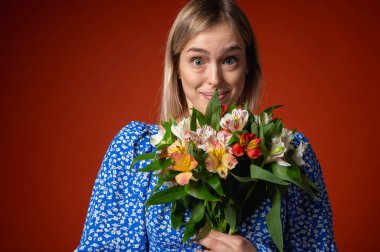 Mavi çiçekli elbisesiyle gülümseyen, güzel bir genç kız elinde parlak turuncu bir arka planda poz veren bir buket çiçek tutuyor. Bahar havası, doğal güzellik, neşeli ruh hali, pozitif