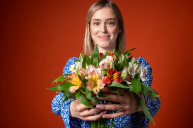Mavi çiçekli elbisesiyle gülümseyen, güzel bir genç kız elinde parlak turuncu bir arka planda poz veren bir buket çiçek tutuyor. Bahar havası, doğal güzellik, neşeli ruh hali, pozitif