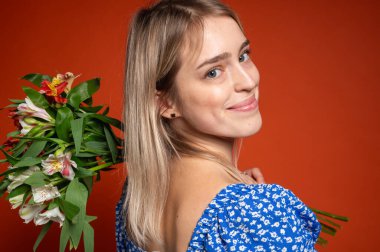 Mavi çiçekli bir elbise giymiş genç bir kadın omuzunda bir buket bahar çiçeği tutuyor, parlak turuncu bir arka planda duruyor. Renkli kontrastlı romantik gizemli poz, beklenti, dikiz açısı