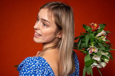 Mavi çiçekli bir elbise giymiş genç bir kadın omuzunda bir buket bahar çiçeği tutuyor, parlak turuncu bir arka planda duruyor. Renkli kontrastlı romantik gizemli poz, beklenti, dikiz açısı