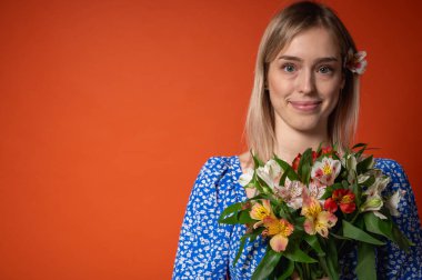 Mavi çiçekli elbisesiyle gülümseyen, güzel bir genç kız elinde parlak turuncu bir arka planda poz veren bir buket çiçek tutuyor. Bahar havası, doğal güzellik, neşeli ruh hali, pozitif