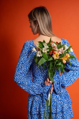 Arkasında parlak turuncu arka planda duran mavi çiçekli bir çiçek buketi tutan genç kadın. Renkli kontrastlı romantik gizemli poz, beklenti