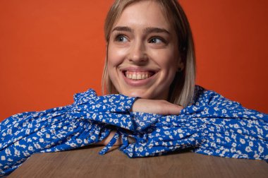 Turuncu arka planda çiçekli mavi elbiseli düşünceli, duygusal genç kız el hareketleriyle. Yüz ifadesi, modern moda ve kendine güvenen duruş, duygular, jestler, yaşam tarzı.