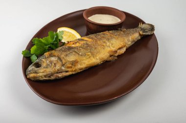 Kahverengi bir tabakta taze maydanoz, limon dilimi ve kremalı sosla servis edilen kızarmış balık. Lezzetli bir deniz ürünleri yemeği, restoran menüsü ya da yemek blogu için mükemmel.