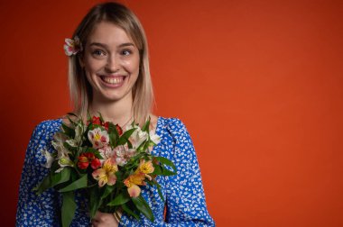 Mavi çiçekli elbisesiyle gülümseyen, güzel bir genç kız elinde parlak turuncu bir arka planda poz veren bir buket çiçek tutuyor. Bahar havası, doğal güzellik, neşeli ruh hali, pozitif