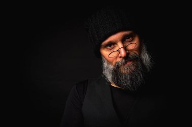 Yuvarlak gözlüklü, siyah örülü şapka ve yelek giyen gri sakallı olgun bir adam. Dramatik ışıklandırması ve karanlık bir geçmişi olan stüdyo portresi. Hippi tarzı, düşünceli, ciddi. Alçak tuş