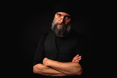 Kolları katlanmış, bere takan ve gözlük takan kendinden emin olgun bir adam. Yoğun bakışlar, gri sakal ve siyah yelek. Koyu arkaplanda gösterişsiz bir portre.