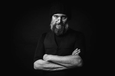 Sert sakallı, örgü şapkalı ve gözlüklü bir adamın yoğun siyah beyaz portresi. Dramatik gölgeler, sade ışıklandırma ve sinema tarzı gizemli bir hava yaratır.