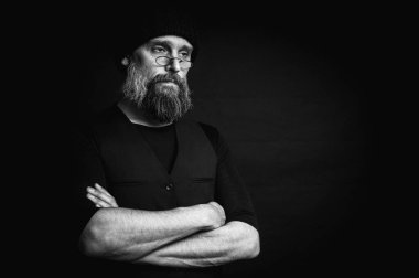 Sert sakallı, örgü şapkalı ve gözlüklü bir adamın yoğun siyah beyaz portresi. Dramatik gölgeler, sade ışıklandırma ve sinema tarzı gizemli bir hava yaratır.