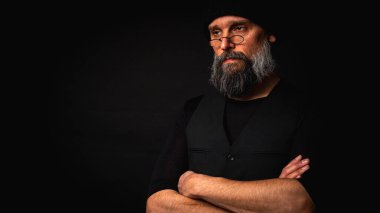 Kolları katlanmış, bere takan ve gözlük takan kendinden emin olgun bir adam. Yoğun bakışlar, gri sakal ve siyah yelek. Koyu arkaplanda gösterişsiz bir portre.