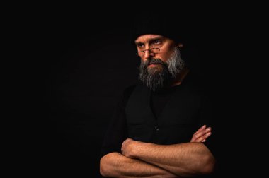 Kolları katlanmış, bere takan ve gözlük takan kendinden emin olgun bir adam. Yoğun bakışlar, gri sakal ve siyah yelek. Koyu arkaplanda gösterişsiz bir portre.