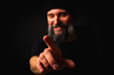 Siyah elbiseli ve şapkalı kızgın sakallı adam kamerayı işaret ediyor. Ciddi bir yüz ifadesi, bastırılmış ışıklandırma. Suçlama, tehdit ya da düzen kavramı. Seçici odaklanma, düşük tuş