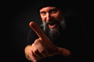 Siyah elbiseli ve şapkalı kızgın sakallı adam kamerayı işaret ediyor. Ciddi bir yüz ifadesi, bastırılmış ışıklandırma. Suçlama, tehdit ya da düzen kavramı. Seçici odaklanma, düşük tuş