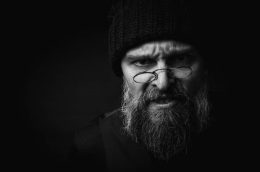Sert sakallı, örgü şapkalı ve gözlüklü bir adamın yoğun siyah beyaz portresi. Dramatik gölgeler, sade ışıklandırma ve sinema tarzı gizemli bir hava yaratır.