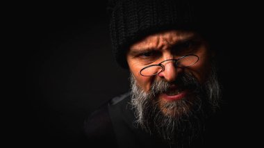 Siyah bereli kızgın sakallı adam ve kameraya homurdanan gözlükler. Gösterişsiz ışıklandırma dramatik portre, anlamlı yüz, duygusal gerilim, karanlık arka plan