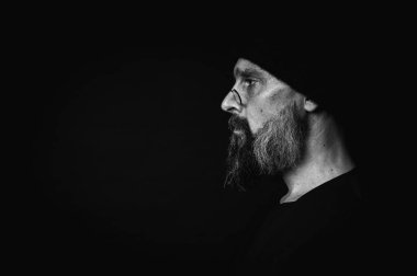 Siyah-beyaz, portre, keskin bakışlı, somurtkan suratlı, gözlüklü, örgü şapkalı, yana bakan etkileyici olgun bir adam. Karanlık arka planda güçlü duyguları olan dramatik bir portre..