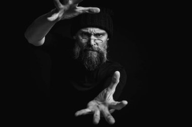 Siyah ve beyaz, kameraya doğru uzanan sakallı ve gözlüklü enerjik bir adamın dramatik portresi. İnce ışıklandırma, duygusal ifade, karanlık arkaplan, gizemli konsept