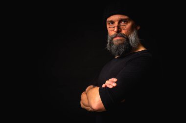 Kollarını önünde kavuşturmuş, kep ve gözlük takan kendinden emin, olgun bir adam. Bakışlar, gri sakal ve siyah yelek. Koyu arkaplanda gösterişsiz, gösterişsiz bir portre..