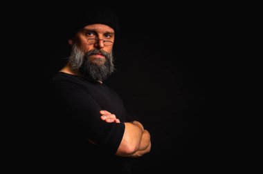 Kollarını önünde kavuşturmuş, kep ve gözlük takan kendinden emin, olgun bir adam. Bakışlar, gri sakal ve siyah yelek. Koyu arkaplanda gösterişsiz, gösterişsiz bir portre..