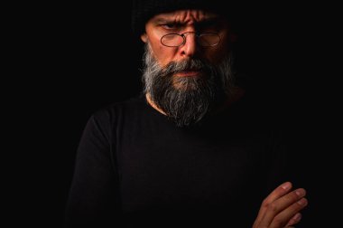 Kollarını önünde kavuşturmuş, kep ve gözlük takan kendinden emin, olgun bir adam. Bakışlar, gri sakal ve siyah yelek. Koyu arkaplanda gösterişsiz, gösterişsiz bir portre..
