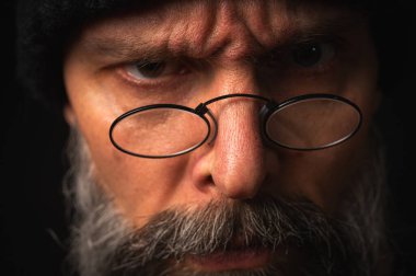 Siyah şapkalı ve kameraya hırlayan gözlüklü kızgın sakallı adamın portresi. Bastırılmış aydınlatma, anlamlı yüz, duygusal gerilim, karanlık arka plan dramatik portre