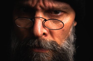 Siyah şapkalı ve kameraya hırlayan gözlüklü kızgın sakallı adamın portresi. Bastırılmış aydınlatma, anlamlı yüz, duygusal gerilim, karanlık arka plan dramatik portre