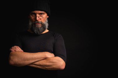 Kollarını önünde kavuşturmuş, kep ve gözlük takan kendinden emin, olgun bir adam. Bakışlar, gri sakal ve siyah yelek. Koyu arkaplanda gösterişsiz, gösterişsiz bir portre..