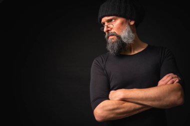 Kollarını önünde kavuşturmuş, kep ve gözlük takan kendinden emin, olgun bir adam. Bakışlar, gri sakal ve siyah yelek. Koyu arkaplanda gösterişsiz, gösterişsiz bir portre..