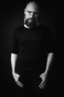 Gri sakallı, yuvarlak gözlüklü, kel bir adamın siyah beyaz portresi hafif bir çimdikleme hareketi yapıyor. Duygular. Siyah arka planda dramatik ışıklandırması ve anlamlı bir yüzü olan sade bir portre..