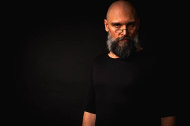 Yoğun bakışlı ve yuvarlak gözlüklü kel sakallı bir adam dramatik bir şekilde hafif ışıklandırılmış. Gizemli görünüş, bilge ifade, karanlık karamsar portre