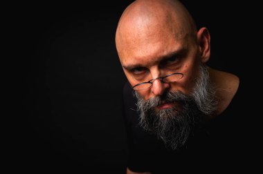 Yoğun bakışlı ve yuvarlak gözlüklü kel sakallı bir adam dramatik bir şekilde hafif ışıklandırılmış. Gizemli görünüş, bilge ifade, karanlık karamsar portre