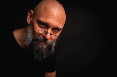 Yoğun bakışlı ve yuvarlak gözlüklü kel sakallı bir adam dramatik bir şekilde hafif ışıklandırılmış. Gizemli görünüş, bilge ifade, karanlık karamsar portre