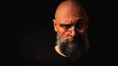 Yoğun bakışlı ve yuvarlak gözlüklü kel sakallı bir adam dramatik bir şekilde hafif ışıklandırılmış. Gizemli görünüş, bilge ifade, karanlık karamsar portre