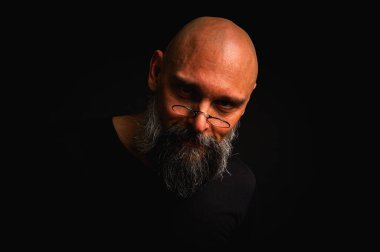 Yoğun bakışlı ve yuvarlak gözlüklü kel sakallı bir adam dramatik bir şekilde hafif ışıklandırılmış. Gizemli görünüş, bilge ifade, karanlık karamsar portre