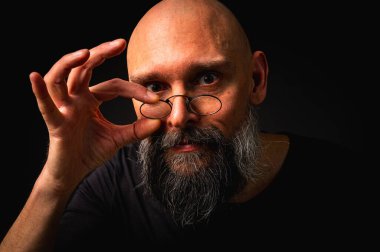 Gri sakallı ve yuvarlak gözlüklü gergin kel bir adam küçük bir çimdik hareketi yapıyor. Dramatik ışıklandırma ve siyah arkaplanda anlamlı yüz ile sade tuş portresi