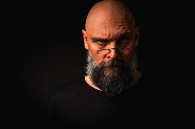 Gri sakallı ve yuvarlak gözlüklü gergin kel bir adam küçük bir çimdik hareketi yapıyor. Dramatik ışıklandırma ve siyah arkaplanda anlamlı yüz ile sade tuş portresi